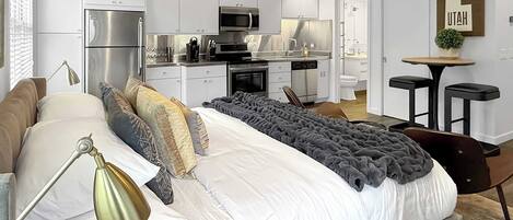 A modern bedroom with a large bed, bedside lamp, and a view into a kitchen area.