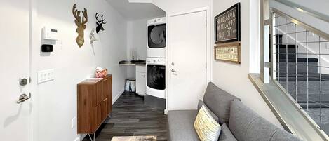 A modern living room with a grey sofa, a wooden cabinet, a washing machine, and a wall-mounted thermostat.