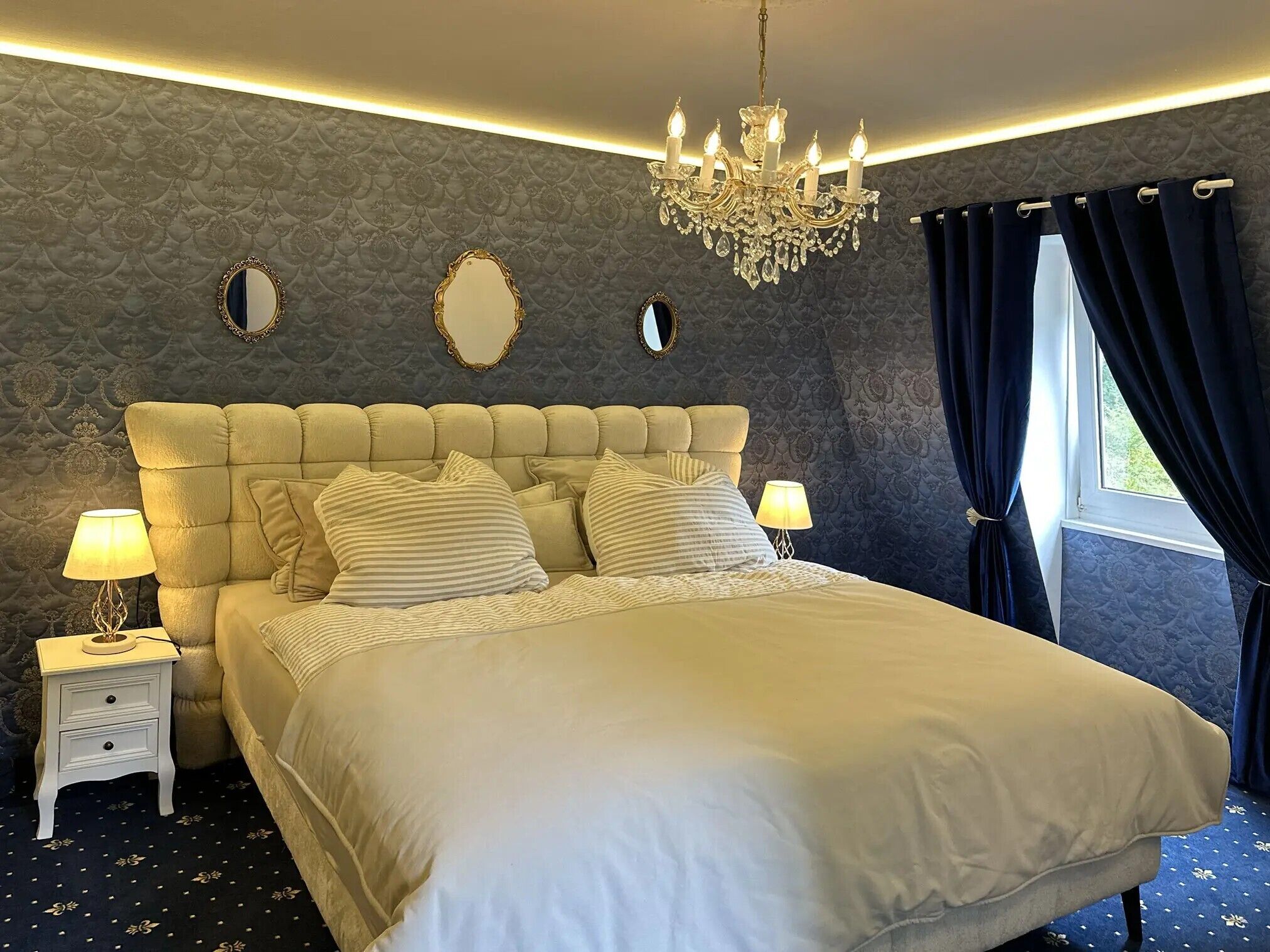 Ein Schlafzimmer mit einem geblümten Kopfteil, zwei Nachttischen mit Lampen, einer Kronleuchter und einem Fenster mit Vorhängen.