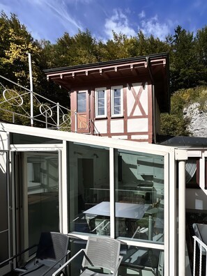 Un chalet doté d’un balcon en verre, de rampes en métal et offrant une vue sur une colline boisée.