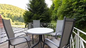 A deck with a round table and four chairs, surrounded by a white railing, overlooking a forested hillside.