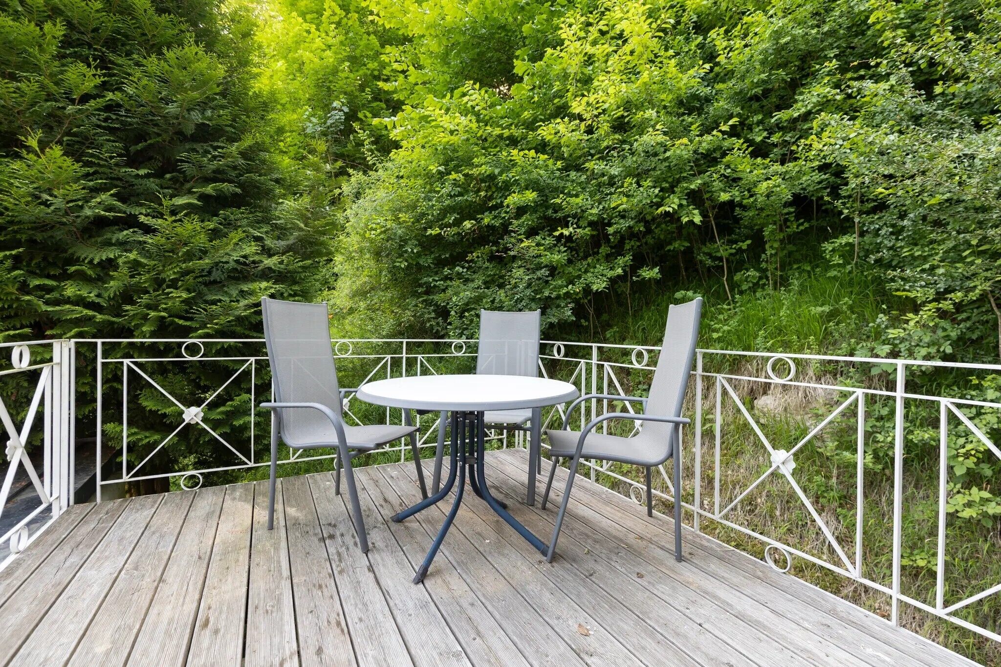Eine Holzterrasse mit einem runden Tisch und zwei Stühlen, umgeben von einem weißen Metallgeländer.