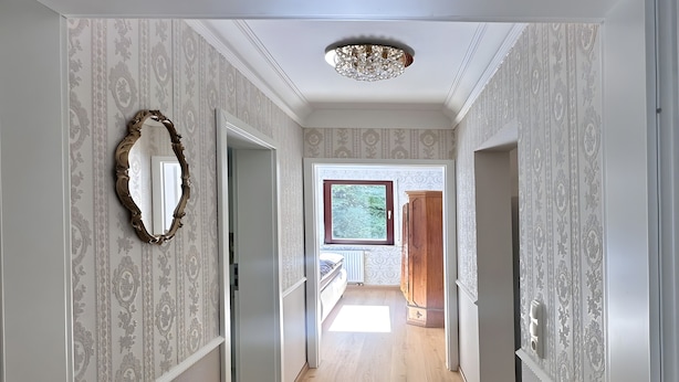 A hallway with patterned wallpaper, wooden flooring, a large mirror, and a chandelier.