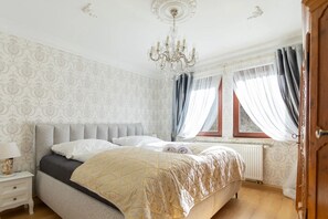 Ein Schlafzimmer mit Bett, Nachttisch, einer Chandelieber, einem Fenster mit Vorhängen und Wandpapier mit Muster.