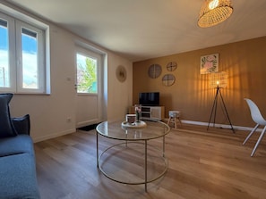 Ein modernes Wohnzimmer mit einem Glastisch, einer Couch, einem Fernseher auf einer Holzkommode, einer Stehlampe und einem großen Fenster mit Blick auf Grünflächen.
