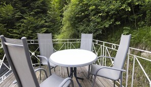 Ein runder Tisch mit vier Stühlen auf einer Holzterrasse, umgeben von einem weißen Metallgeländer, mit dichtem Grün im Hintergrund.