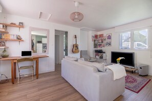 Ein Wohnzimmer mit einer Couch, einem Schreibtisch mit Laptop, einer Gitarre, einem Fernseher und einem Bücherregal.