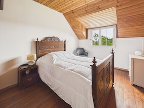 Ein Schlafzimmer mit Holzbalkendecke, einem Bett mit Kopfteil, einem Nachttisch, einem Stuhl und einem Fenster.