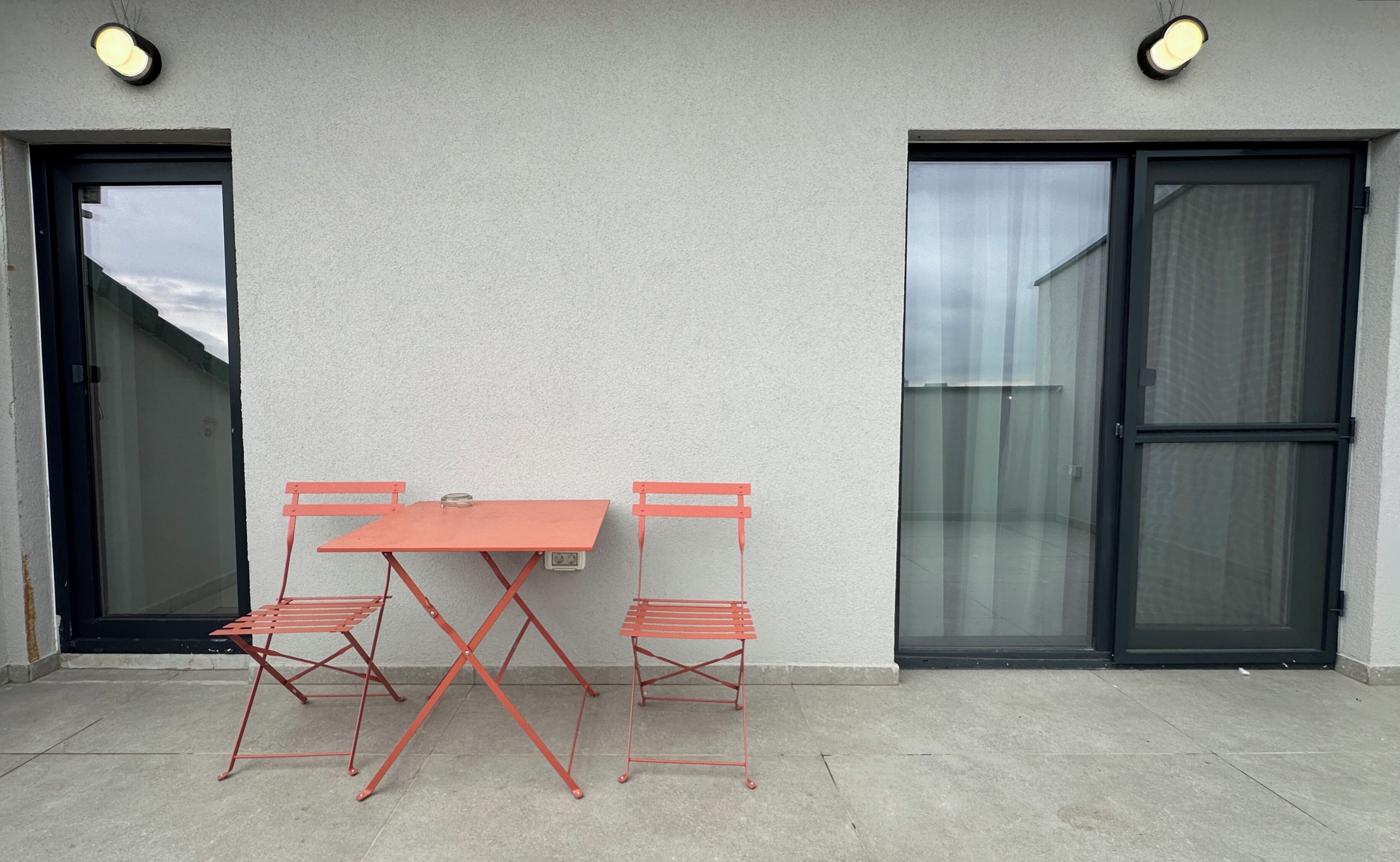 Een terras met een tafel en twee stoelen, een schuifpui en een wandlamp.