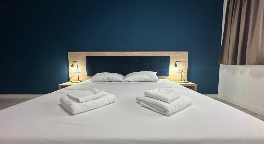 Quarto de hotel com cama, duas luminárias laterais e toalhas dobradas na cama.