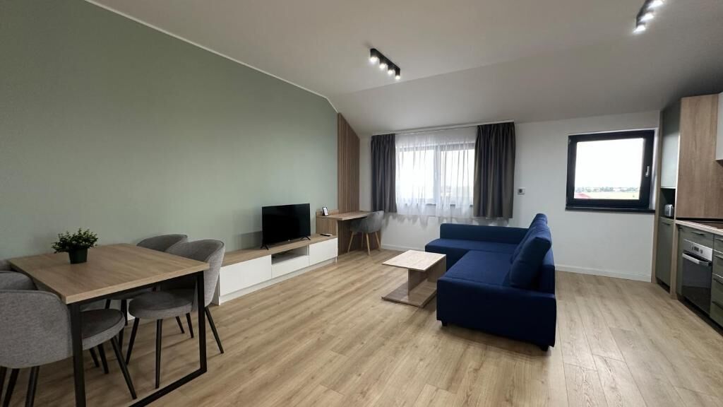 A modern living room with a sofa, a dining table with chairs, a TV, and a kitchen area with cabinets and a window.