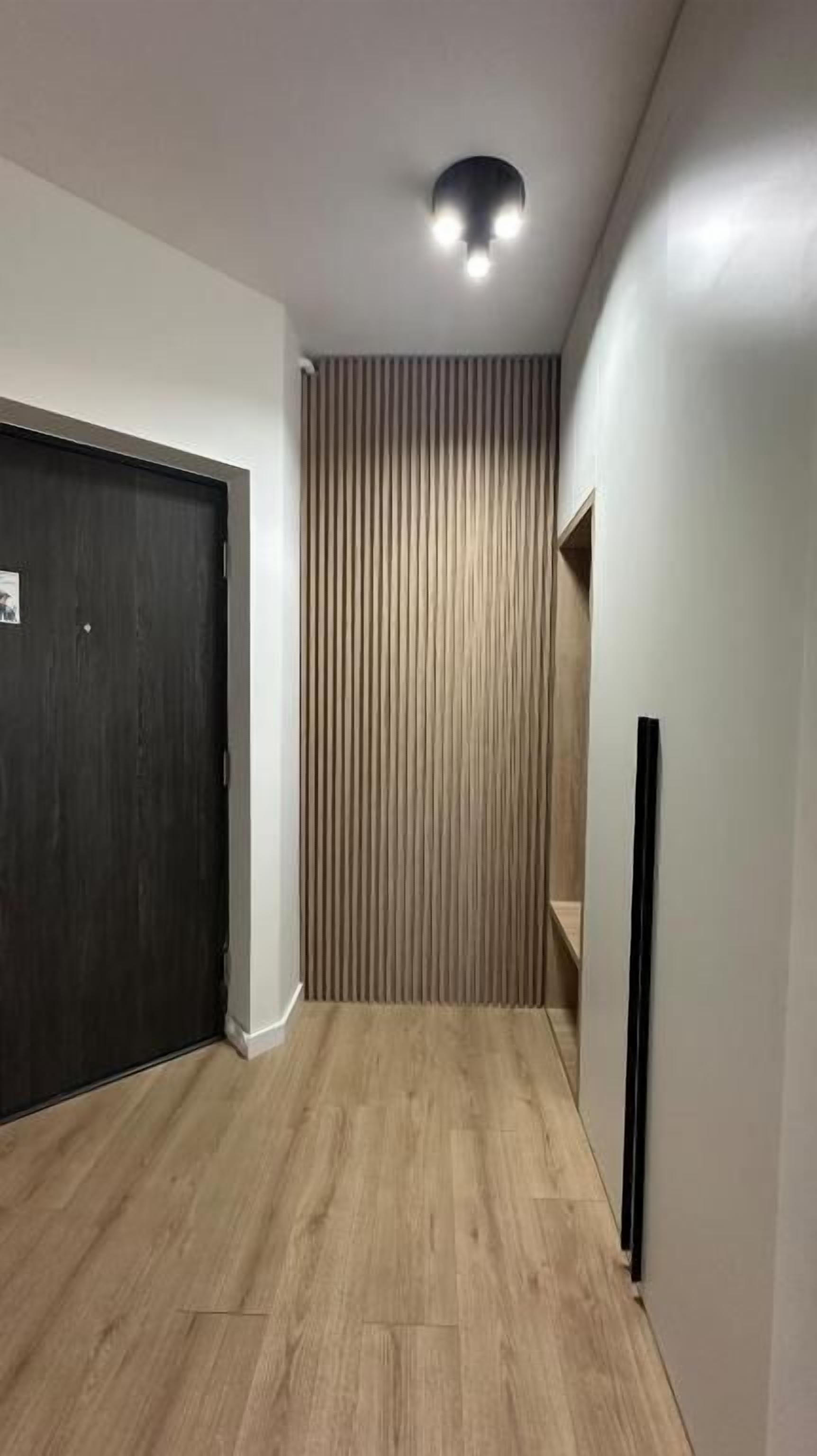 Un pasillo con una puerta oscura, listones de madera verticales, una lámpara y un objeto negro en la pared.