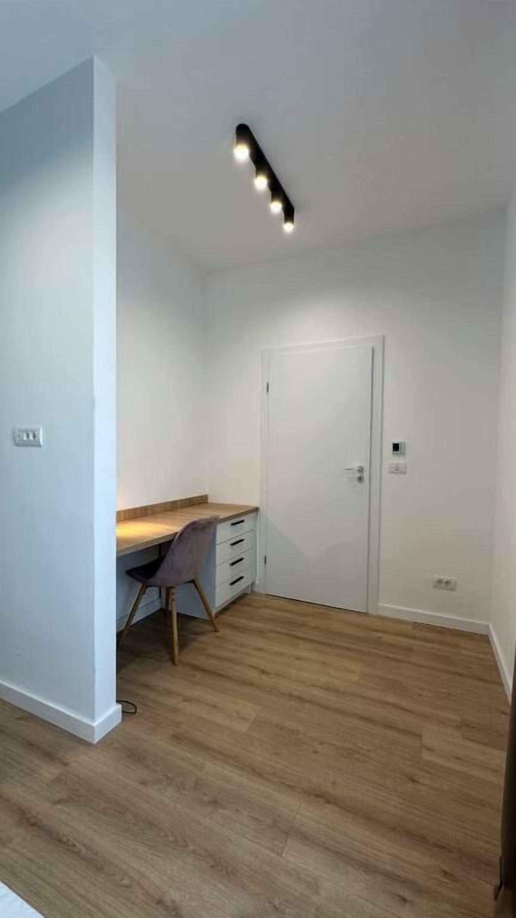 Una habitación con piso de madera, un escritorio con silla, una puerta blanca y una lámpara en el techo.