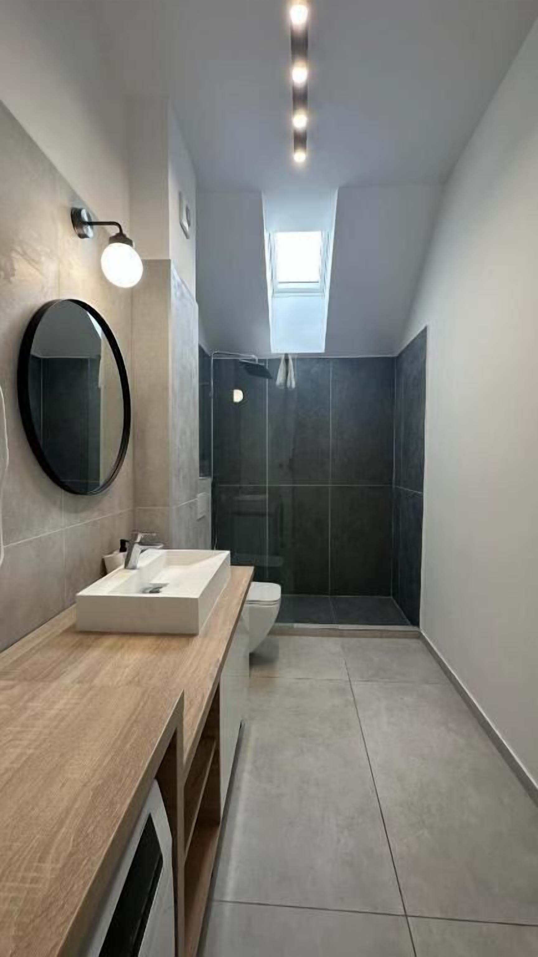 A modern bathroom with a wooden vanity, a round mirror, a shower area, and a skylight.