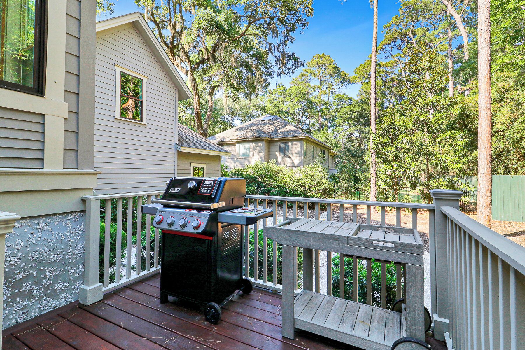 A deck with a grill, a house in the background, and surrounding trees.