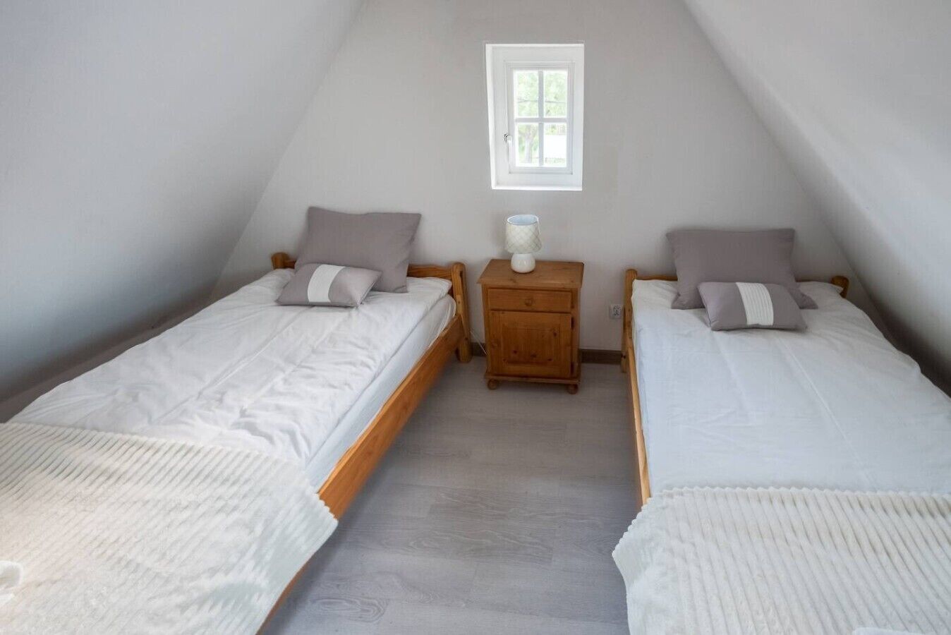 Quarto com duas camas de solteiro, uma mesinha de cabeceira de madeira, uma janela e um abajur branco.