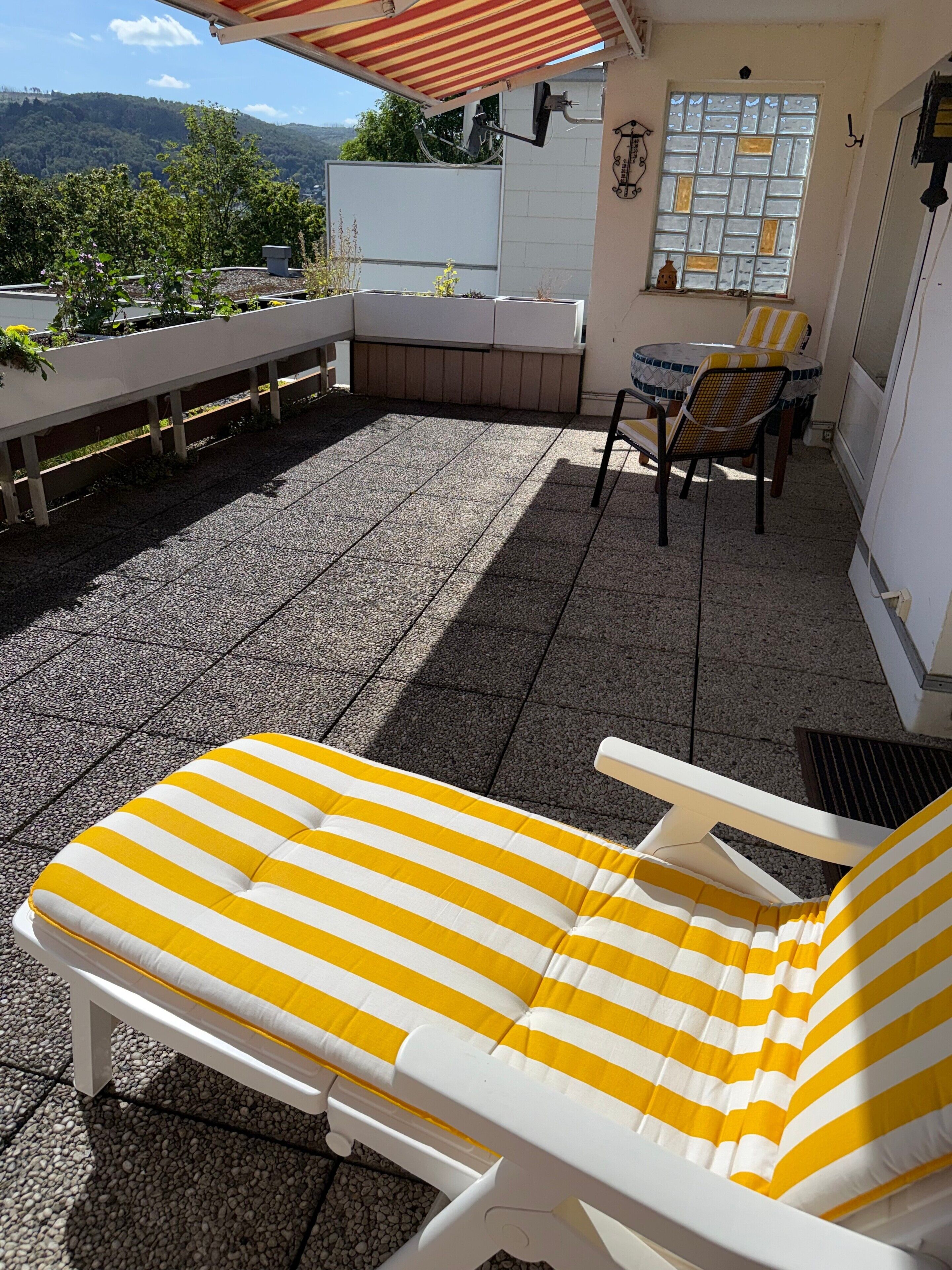 Een zonnig terras met een lounge stoel en een tafel, omringd door een wit hek en uitzicht op een heuvel.