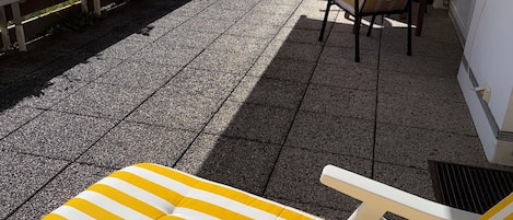 A sunlit patio with a lounge chair and a table, surrounded by a white fence and a view of a hill.