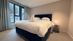A modern bedroom with a large bed, a bedside table with a lamp, and a view of trees through the window. - The Ironworks (Ulverston)