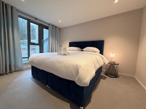 Um quarto moderno com uma cama grande, uma mesa de cabeceira com abajur e vista para árvores pela janela.