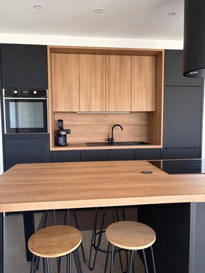 A modern kitchen with wooden cabinets, a built-in oven, a sink, and two stools. - House with a garden on the French-Swiss border – 5 minutes from Geneva (Collonges-sous-Salève)