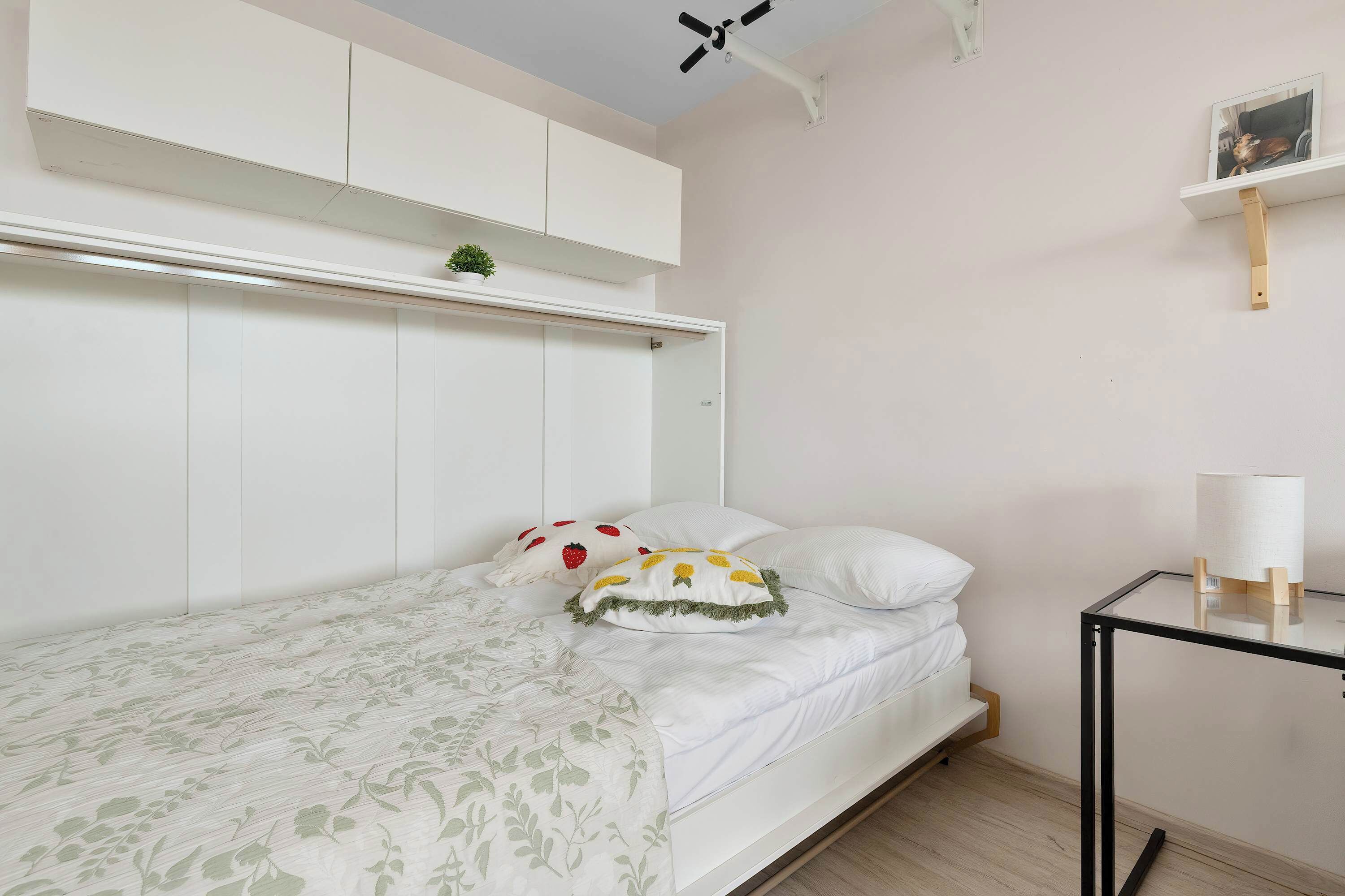 A bedroom with a bed, a bedside table, a wall-mounted shelf with a plant, and a ceiling fan.