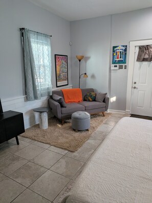 Quarto com sofá cinza, um banquinho redondo pequeno, cama branca, mesa de cabeceira preta, tapete bege, luminária de chão e porta com um quadro em cima.