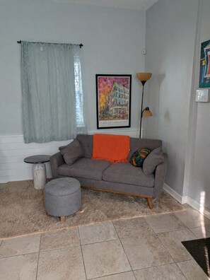 Uma sala de estar com um sofá cinza, um banquinho, um quadro emoldurado, um abajur e uma janela com cortina.