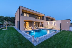 Et moderne hus med to etager, swimmingpool, udendørs møbler og en stenvæg.
