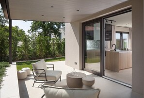 Eine moderne Außenpatio mit minimalistischem Design, mit einer Sitzecke inklusive Sofa und Sesseln, einem runden Tisch und einer Glastür, die zu einer Küche führt.