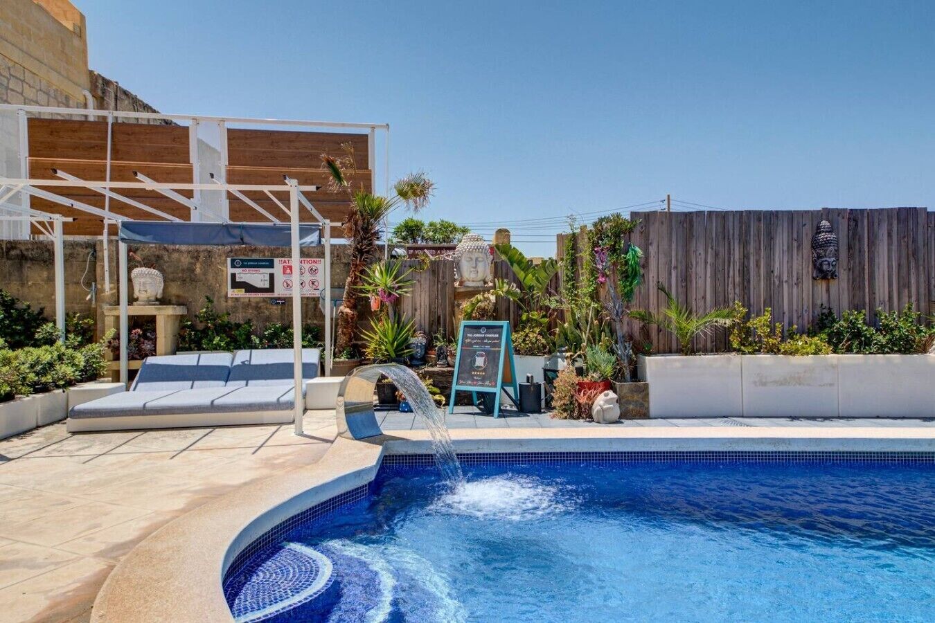 A pool area with a wooden deck, a lounge chair, a small waterfall feature, and a signboard.