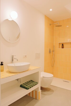 Casa de banho com lavatório branco, espelho e sanita, chuveiro com azulejos amarelos e prateleira com artigos de higiene pessoal.