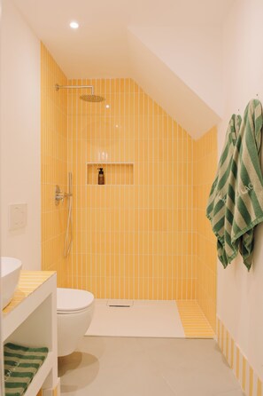 A bathroom with yellow-striped tiles, a white toilet, a shower with a handheld showerhead, and a towel rack with a green towel. - Cute studio in a tiny house right next to the lake (Sala Comacina)