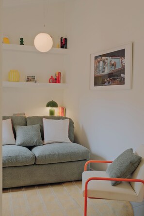 Uma sala de estar com sofá, poltrona, luminária, quadro e prateleiras com diversos objetos.