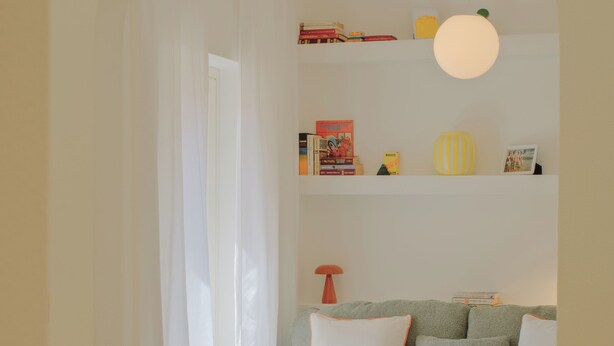 Habitació amb un sofà, una catifa rodona teixida, una cadira vermella i una prestatgeria plena de llibres i objectes decoratius.