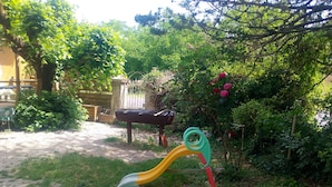 A playground with a slide and swings, surrounded by greenery and a stone wall.