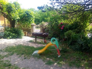 Un parque infantil con tobogán y columpios, rodeado de follaje y un muro de piedra.