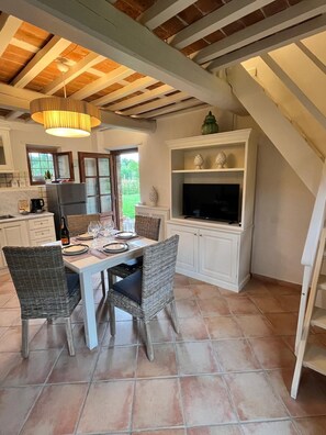 A dining area with a table set for four, surrounded by wicker chairs, a white cabinet with decorative items, and a television on a shelf. - View of Castelvecchio exclusive Tuscan guest house in the countryside of Lucca (Castelvecchio di Cómpito)