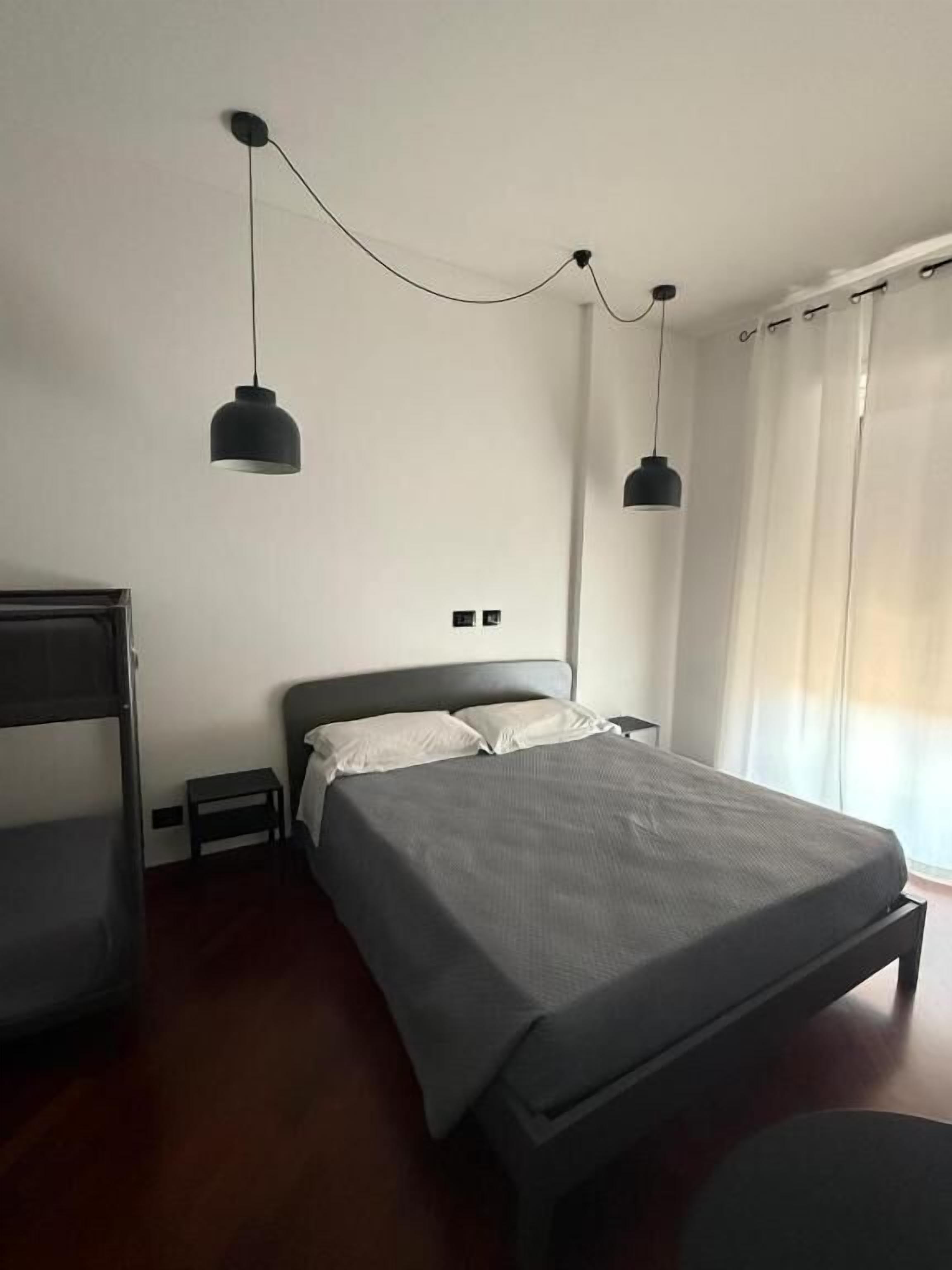 A bedroom with a bed, two pendant lights, a nightstand, a chair, and a window with curtains.