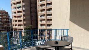 Ein Balkon mit rundem Tisch und Stühlen, einem blauen Geländer und Hochhäusern im Hintergrund.