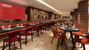 A modern restaurant with long communal tables and booths, a wall of decorative masks, and a red sign with the hotel name.