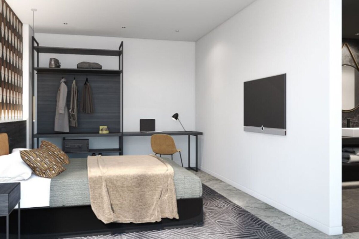 Un dormitorio moderno con cama, escritorio y silla. Hay un armario con ropa colgada y un televisor de pantalla plana montado en la pared.