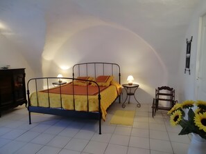 Un dormitori amb un llit de metall, dues taules de nit amb làmpades, una cadira de fusta i un vas amb girassols.