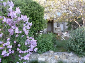 Un mur de pierres, une chaise pliante blanche et une table dans un jardin où poussent des fleurs violettes.