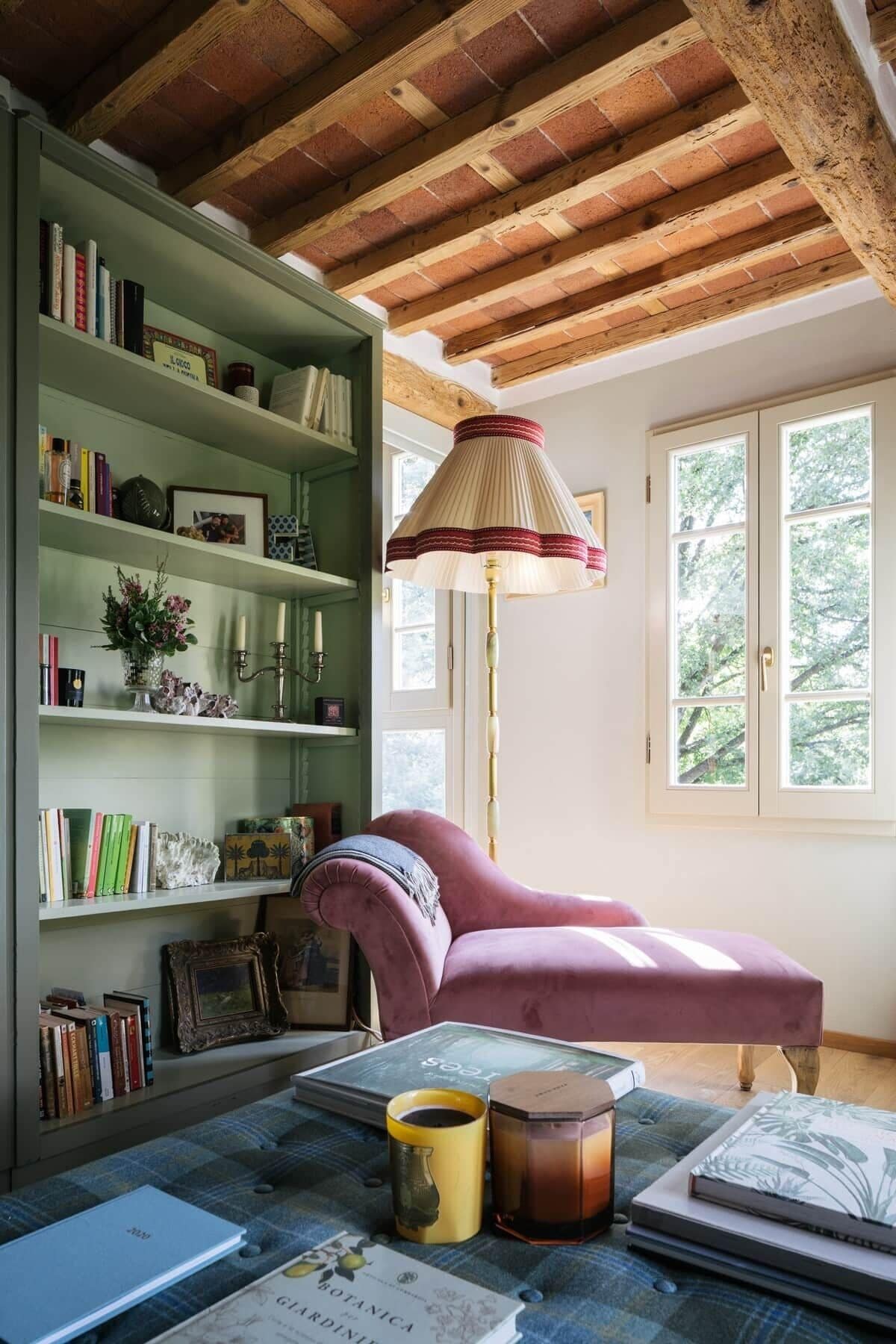 Une pièce avec une bibliothèque verte, un fauteuil rose, une lampe sur pied et une fenêtre donnant sur des arbres.
