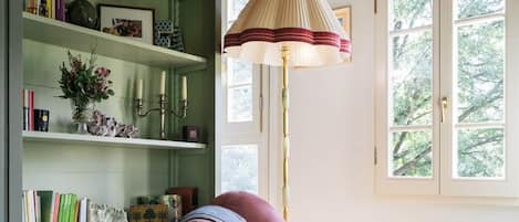 Une pièce avec une bibliothèque verte, un fauteuil rose, une lampe sur pied et une fenêtre donnant sur des arbres.