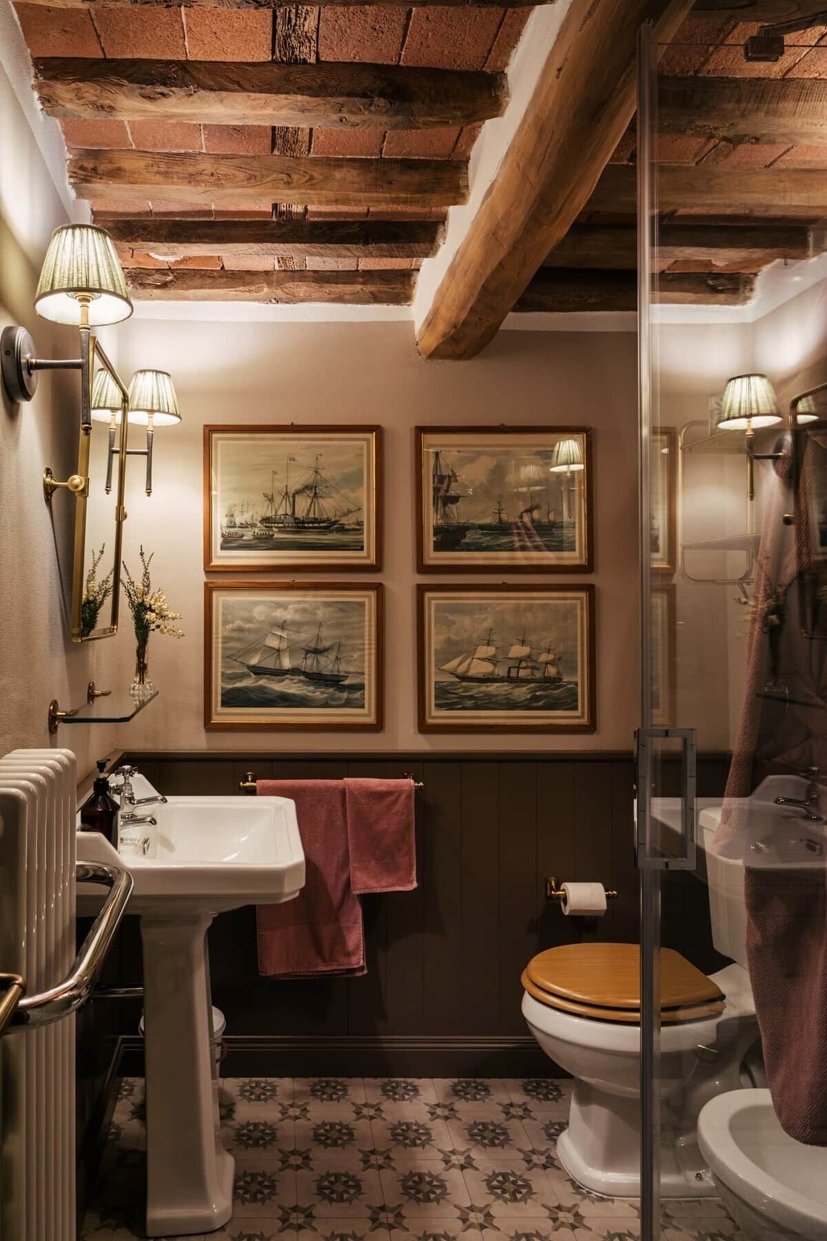 Une salle de bain avec un plafond en bois, un lavabo blanc, des toilettes en bois et quatre tableaux marins encadrés accrochés au mur.
