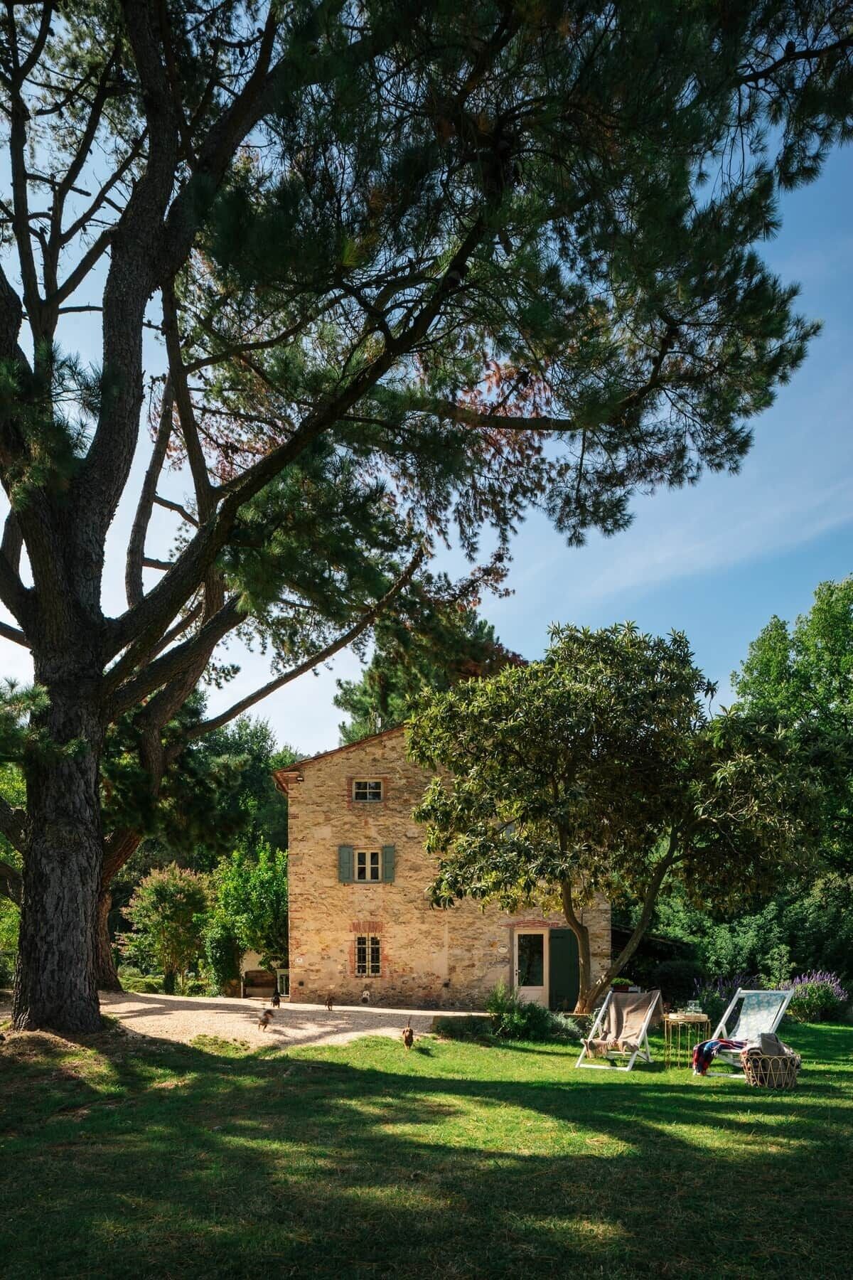 A stone building with a large tree in front, a grassy area, and two lounge chairs.