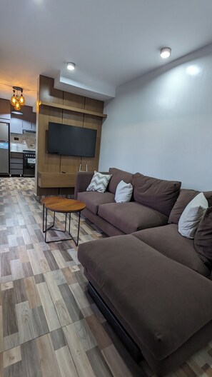 A modern living room with a brown sofa, a small wooden table, and a flat-screen TV mounted on a wooden panel wall. - Cozy One bedroom apartment in Kigali , Rebero with panoramic mountain views (Kigali)