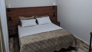Une chambre à coucher avec une tête de lit en bois, un lit recouvert d’une courtepointe, une table de chevet, une lampe fixée au mur et un luminaire au plafond.